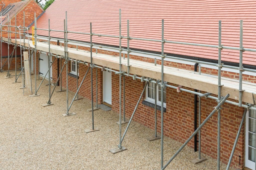 A brick building with a sloped red tile roof is under renovation. Metal scaffolding runs along the side, elevated on a gravel ground. The scaffolding includes a wooden walkway at roof level.