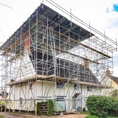 A house surrounded by an extensive scaffolding structure for renovation. The scaffolding is metal, covering the entire roof and extending to the sides. The sky is partly cloudy, and a hedge is visible in the foreground.