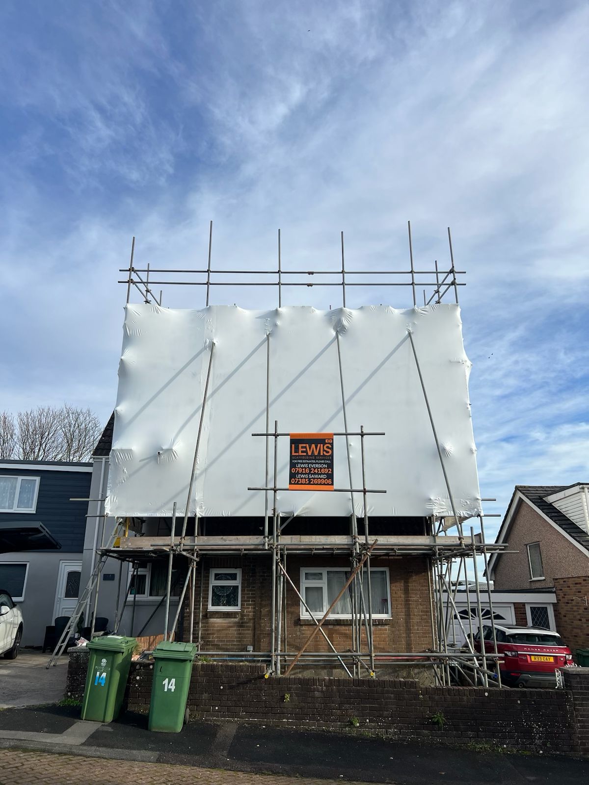 A house under construction is covered with white scaffolding. A sign with "LEWIS" and contact details is displayed on the front. Two green bins are on the left, and a red car is partially visible on the right. The sky is clear and blue.