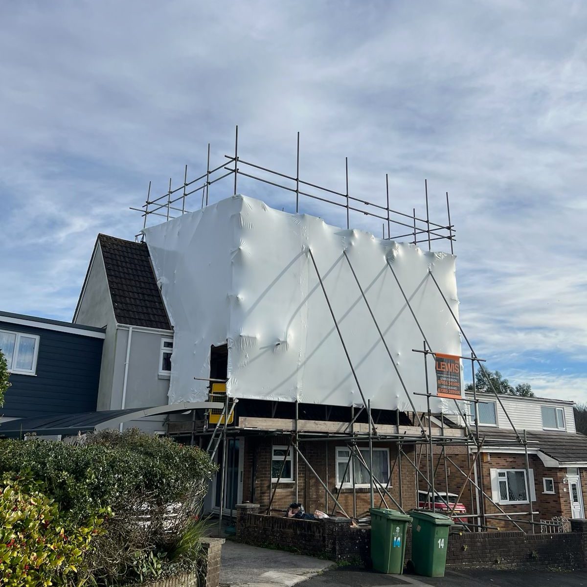 A house covered in white protective sheeting, surrounded by scaffolding. The sky is partly cloudy. Two green trash bins are near the driveway, and there are neighboring houses visible in the background.