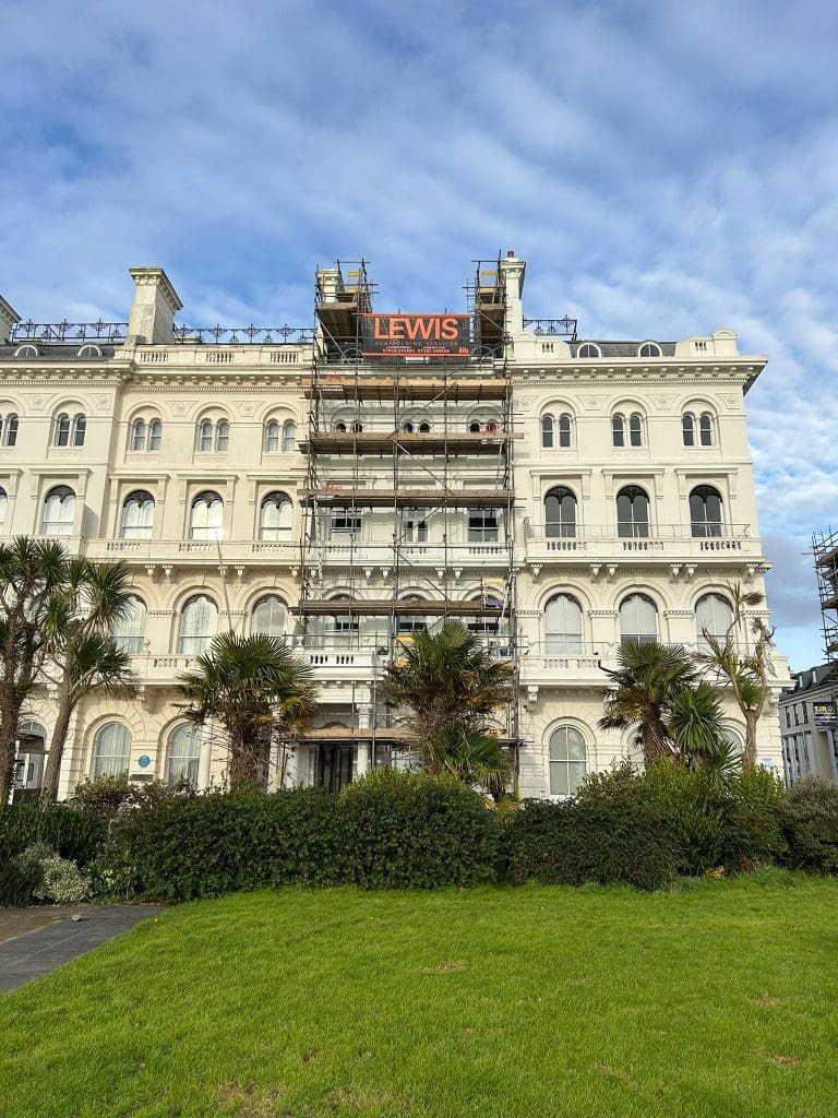 A large white building with classic architectural details is partially covered in scaffolding, with a sign displaying "LEWIS" at the top. Palm trees and a green lawn are in the foreground, under a blue, partly cloudy sky.