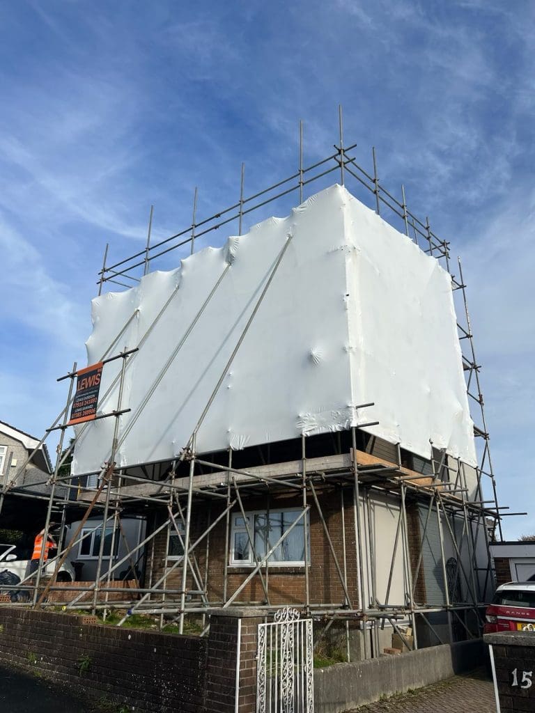 A two-story house is covered in white protective material, surrounded by a metal scaffold. A sign on the scaffolding reads "LEWIS." The house is located on a street with a brick wall and gate at the front.