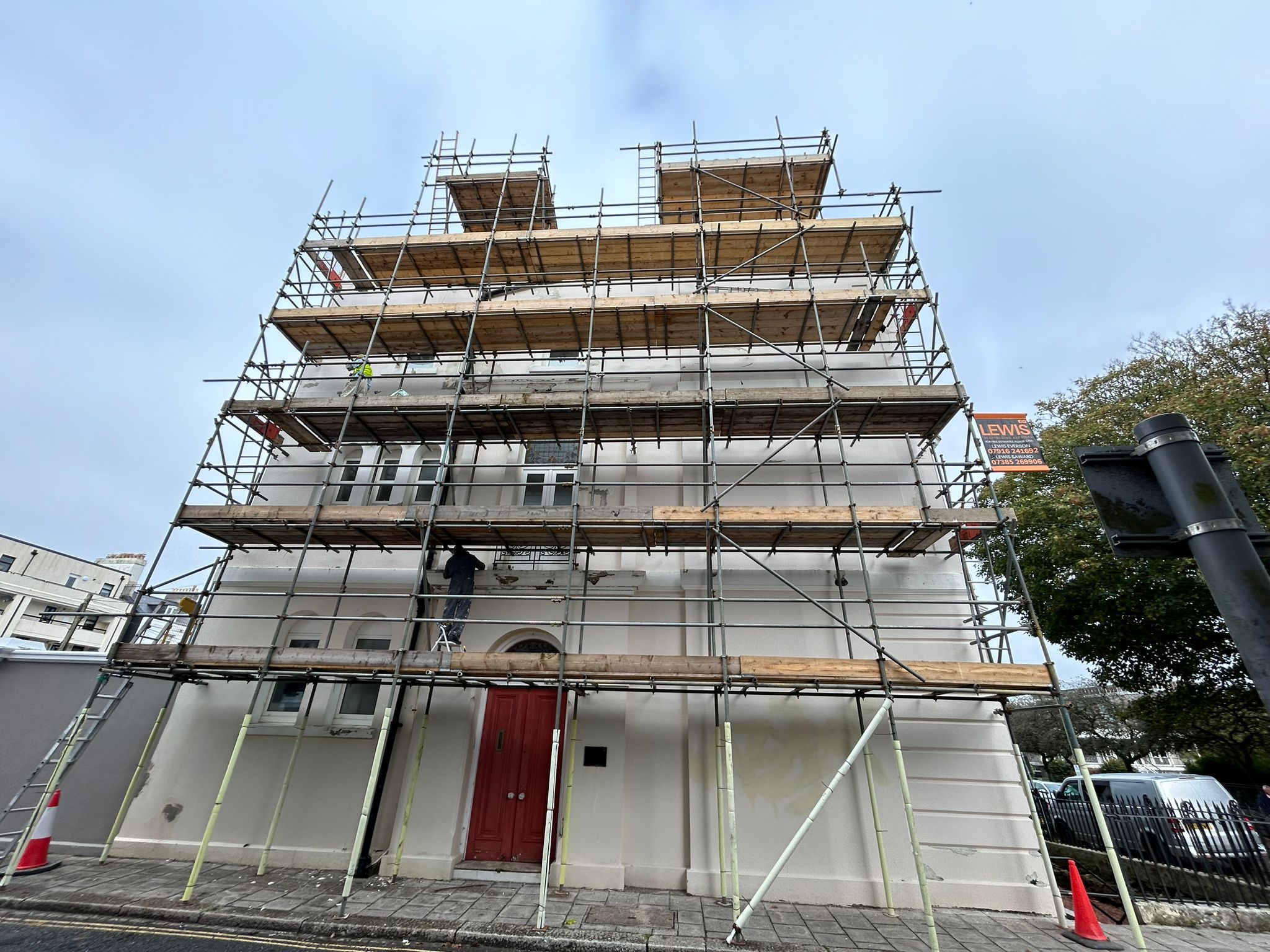 A building covered in scaffolding under a clear sky, with one person working on it. The structure has multiple levels for access. Orange cones are placed on the sidewalk near the site.