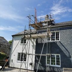 A two-story house with scaffolding set up on the roof and side. The exterior is partially white and partially gray. A ladder leans against the scaffolding, and the sky is clear and blue. A stone path leads up to the building.