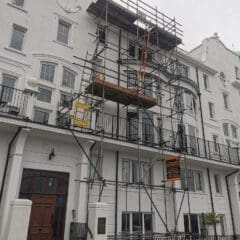 A white multi-story building with scaffolding in front. The scaffolding has a "Lewis" construction sign. The building features multiple windows and a dark brown door.