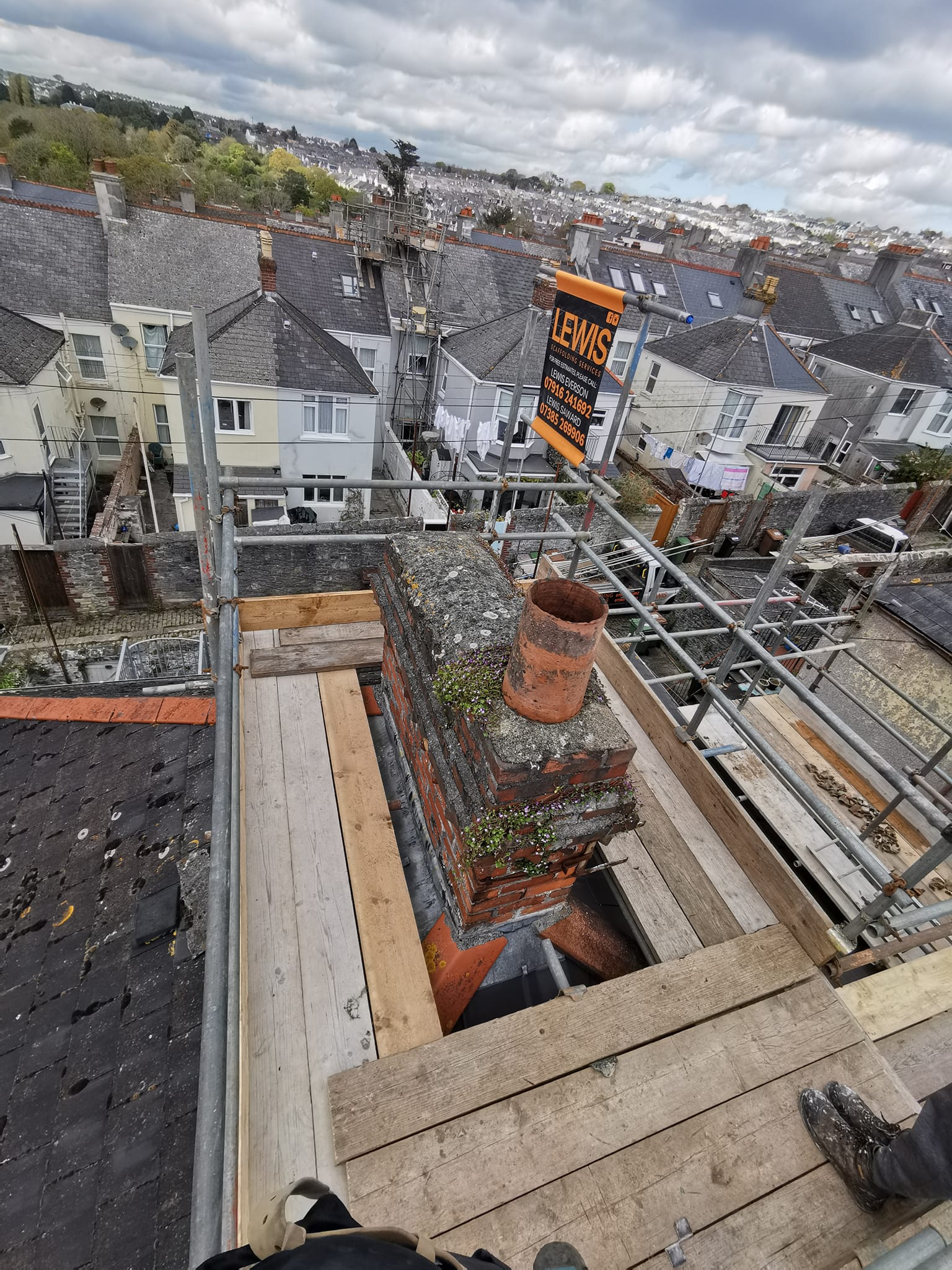 A view from a rooftop shows scaffolding surrounding a brick chimney. Weathered houses and a cloudy sky are in the background. A sign on the scaffolding reads "LEWIS," suggesting construction or renovation work. Planks are laid around the chimney.