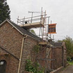 Scaffolding on a stone building with a sign for "Lewis" construction company, displaying contact numbers. An antenna is visible on the roof. The scene includes greenery and a cloudy sky.