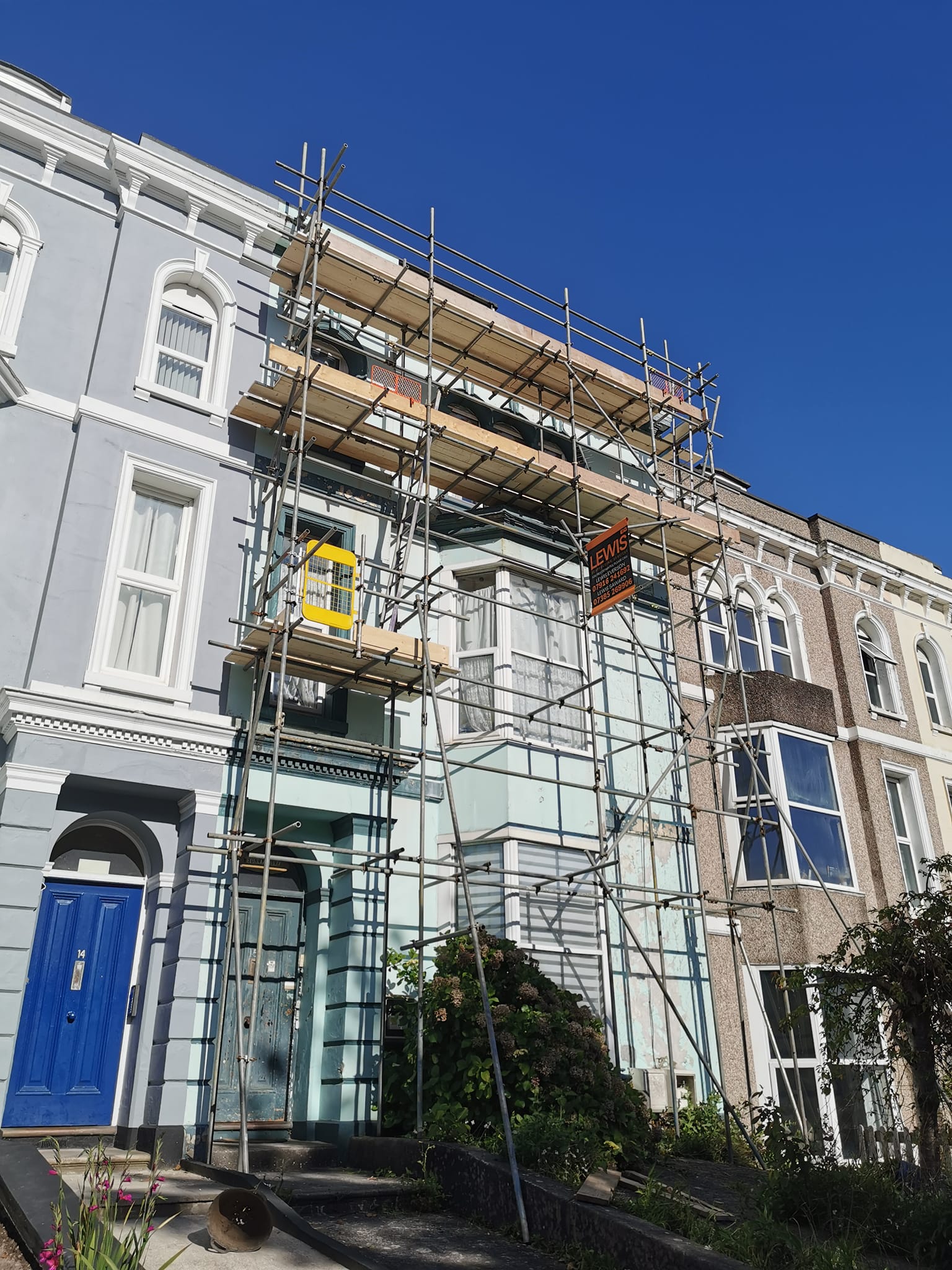 A row of Victorian-style houses with one covered in scaffolding on a bright day. The house under construction has a sign on the scaffolding. A blue door is visible below the structure, and the sky is clear and blue.