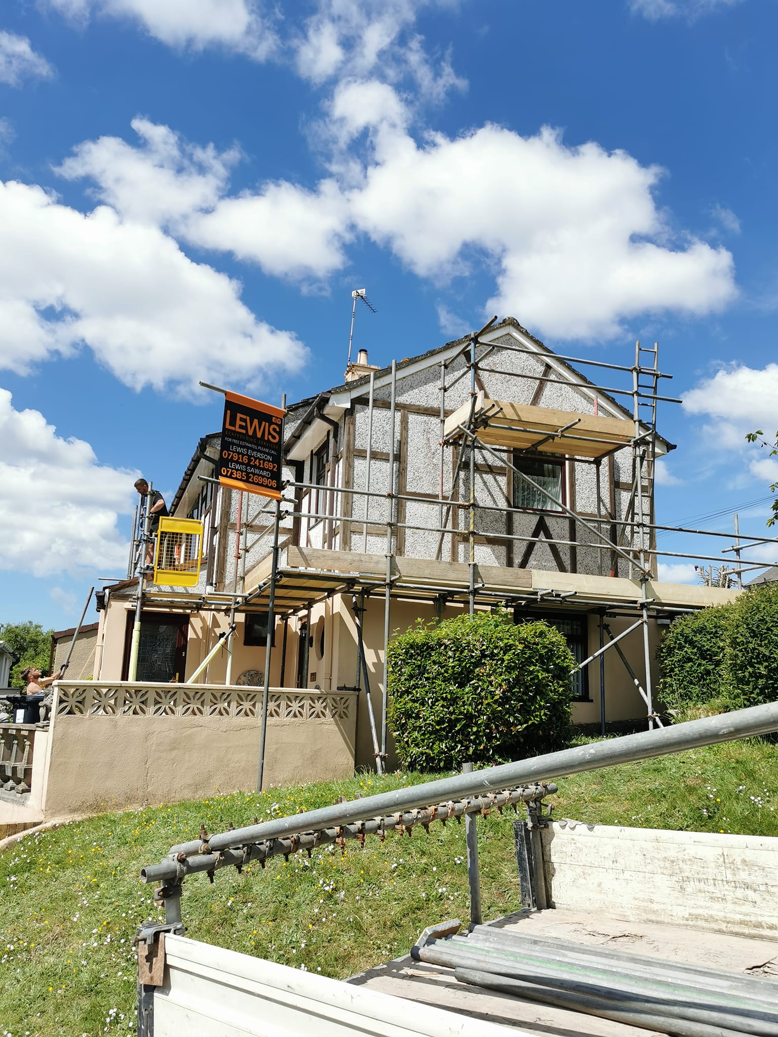 A house under renovation with scaffolding around it. A sign reads "LEWIS." The sky is bright blue with scattered clouds. A grassy slope and a portion of a fence are visible in the foreground.