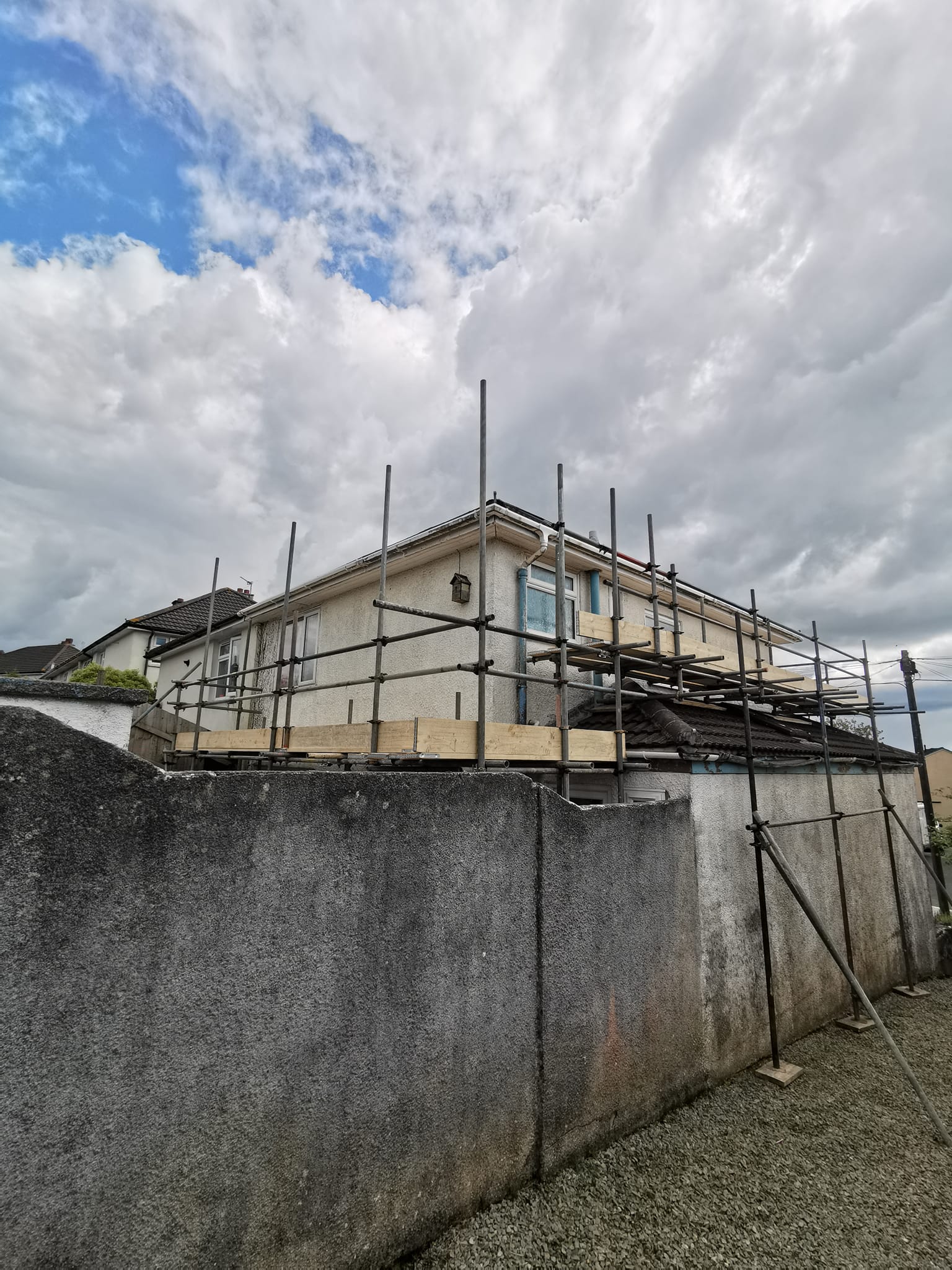 A house under renovation with scaffolding surrounding its exterior. The sky is cloudy, and a concrete wall is visible in the foreground, with a gravel surface below.