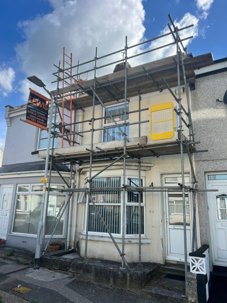 A two-story house with scaffolding set up on the facade for renovation. A sign on the scaffolding reads "Lewis." The neighboring houses are visible, and the sky is partly cloudy.