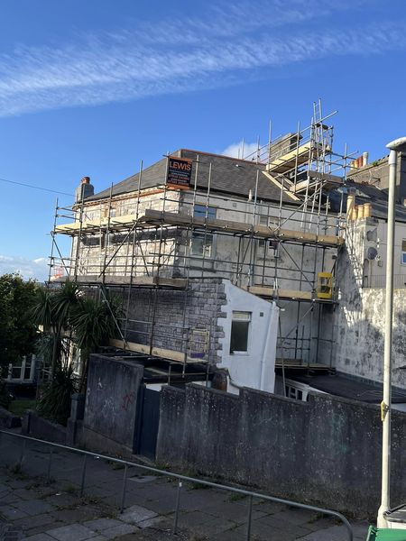 A house undergoing construction, surrounded by scaffolding. The sky is clear and blue, and there are a few clouds. Nearby, there is a small tree and a paved walkway.