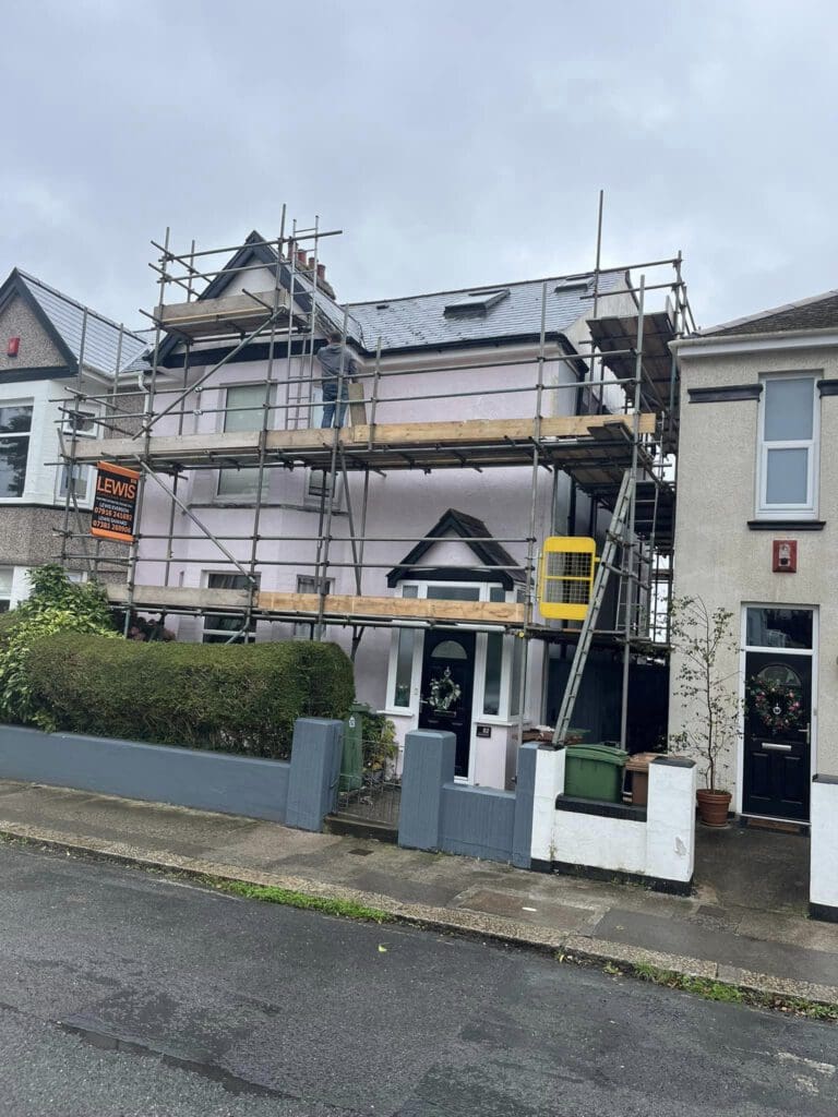 A two-story house under construction or renovation, surrounded by scaffolding. The house has a light pink exterior with a roofed porch. It is located on a residential street with neighboring homes adjacent to it. The sky is overcast.