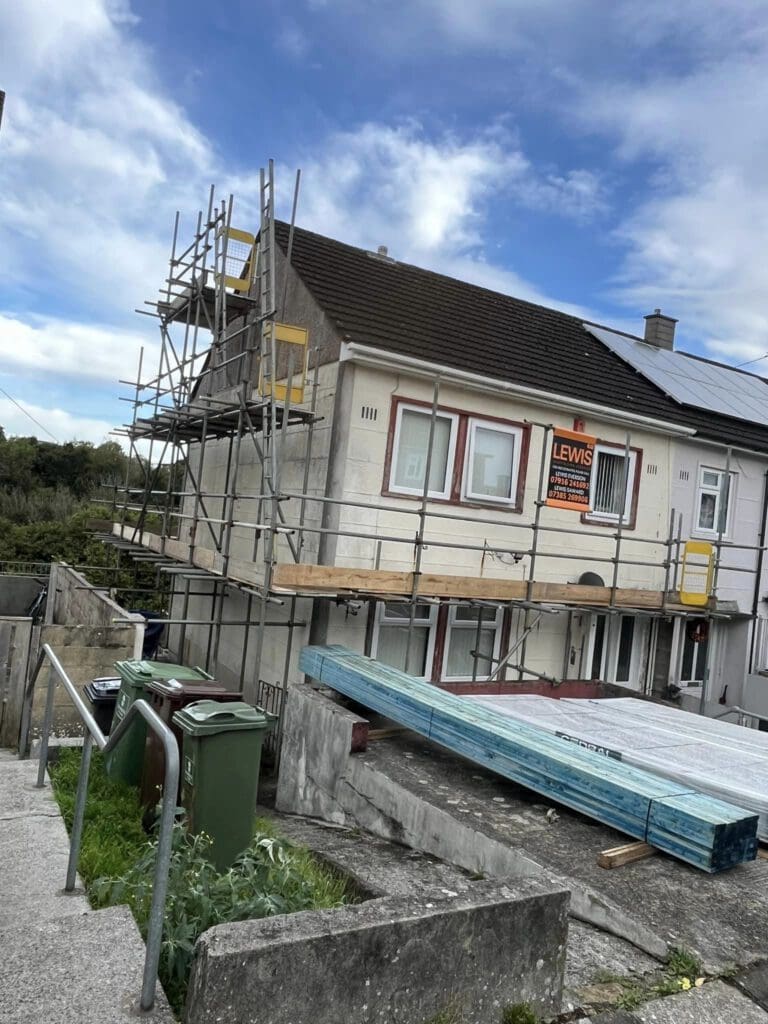 A house under renovation with scaffolding on the exterior. Windows are covered with wooden boards. Building materials and wooden planks are on the ground. A sign with text is visible on the wall. Nearby, green garbage bins are placed by a stairway.