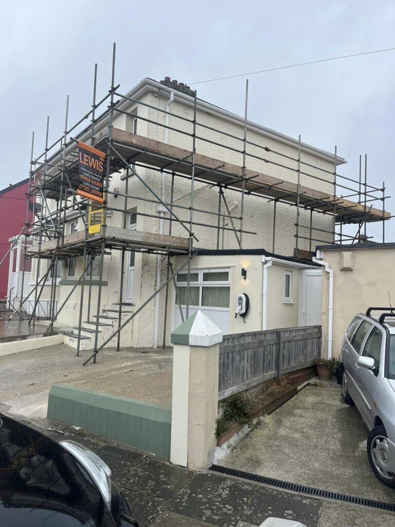 A two-story house is surrounded by scaffolding, indicating ongoing construction or renovation. The house has a beige exterior, and several cars are parked in the driveway and street. A sign is attached to the scaffolding.