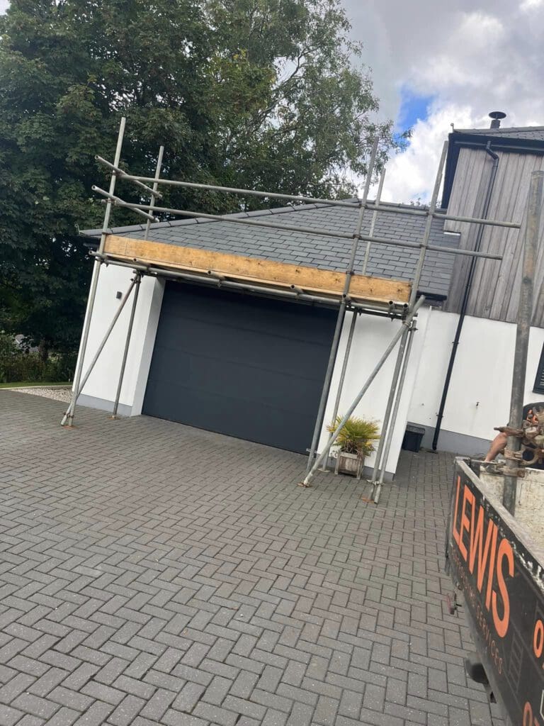 A garage with a dark gray door is surrounded by scaffolding, indicating ongoing construction or maintenance. The foreground shows a paved driveway with a potted plant to the right. Trees are visible in the background under a partly cloudy sky.