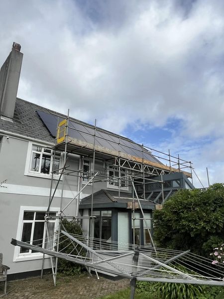 A two-story house with scaffolding erected along one side, wooden boards on the structure. Solar panels are installed on the roof. The sky is cloudy, and there's a rotary clothesline in the foreground with shrubs on the right.