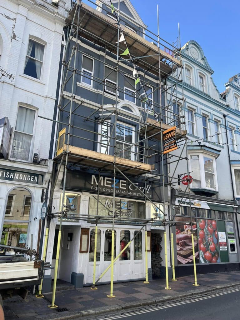 A building under construction with a scaffolding framework covers a restaurant named "Meze Grill." The sign mentions "Greek & Mediterranean Restaurant." Adjacent buildings are visible, and there is a clear blue sky above.