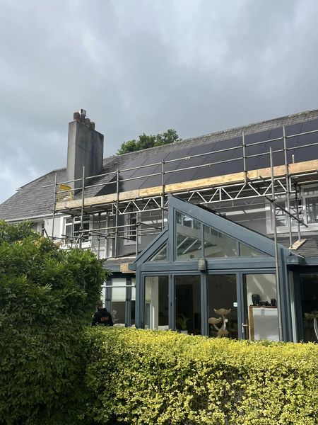 A house with scaffolding on the roof, featuring solar panels being installed. A glass conservatory extends from the house, and a green hedge surrounds the area. The sky is cloudy.