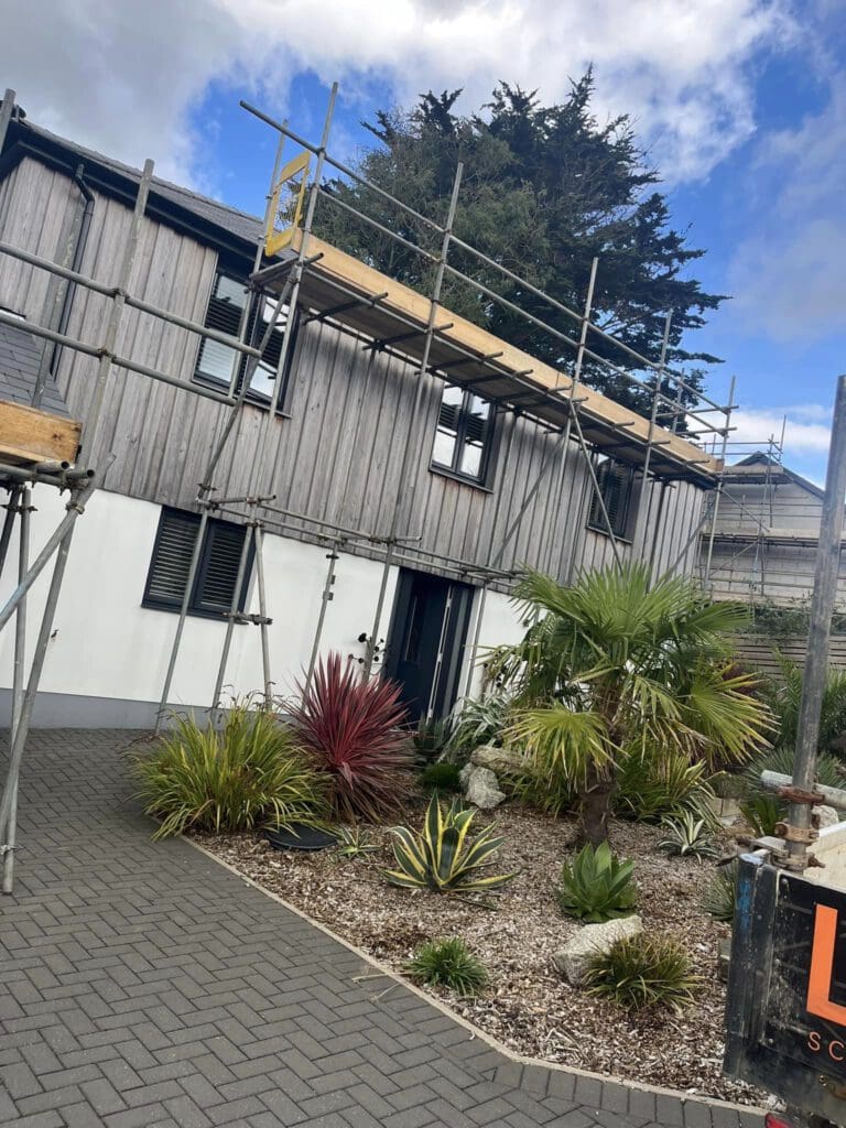 A two-story house with wooden cladding is under renovation, featuring scaffolding on the exterior. There is a garden in front with various plants and shrubs. The sky is partly cloudy.