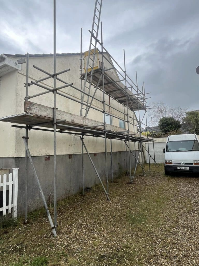 A house under construction with scaffolding on its side. A white van is parked nearby on a gravel driveway. The sky is overcast, suggesting cloudy weather.