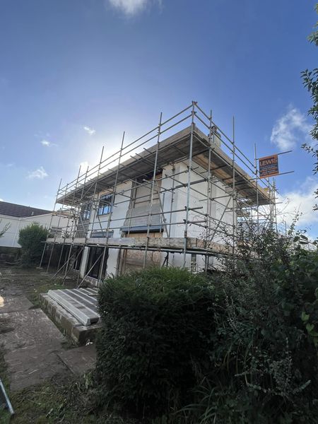 A two-story house under renovation with scaffolding surrounding it. The sky is clear with some clouds, and there are construction materials on the ground. Bushes and greenery are visible in the foreground.