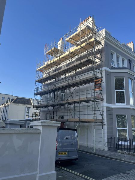 A four-story building under renovation with scaffolding covering its facade. A grey van parked in front and a clear blue sky overhead.