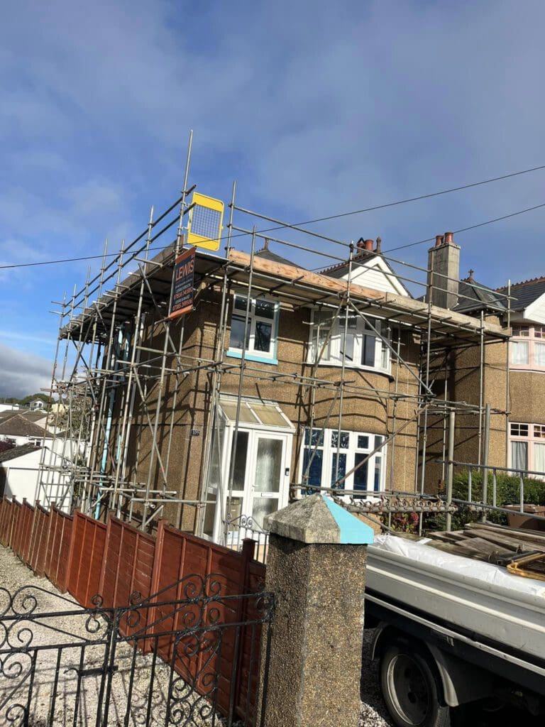 A two-story house with scaffolding around it, a sign labeled "LEWIS" is attached to the scaffolding. The sky is partly cloudy, and a gate and a truck are visible in the foreground.