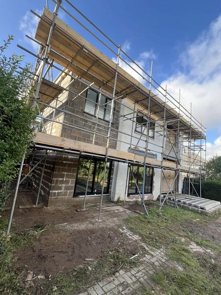 A two-story house under construction with scaffolding surrounding it. The building is made of concrete blocks, and there is a clear blue sky in the background.