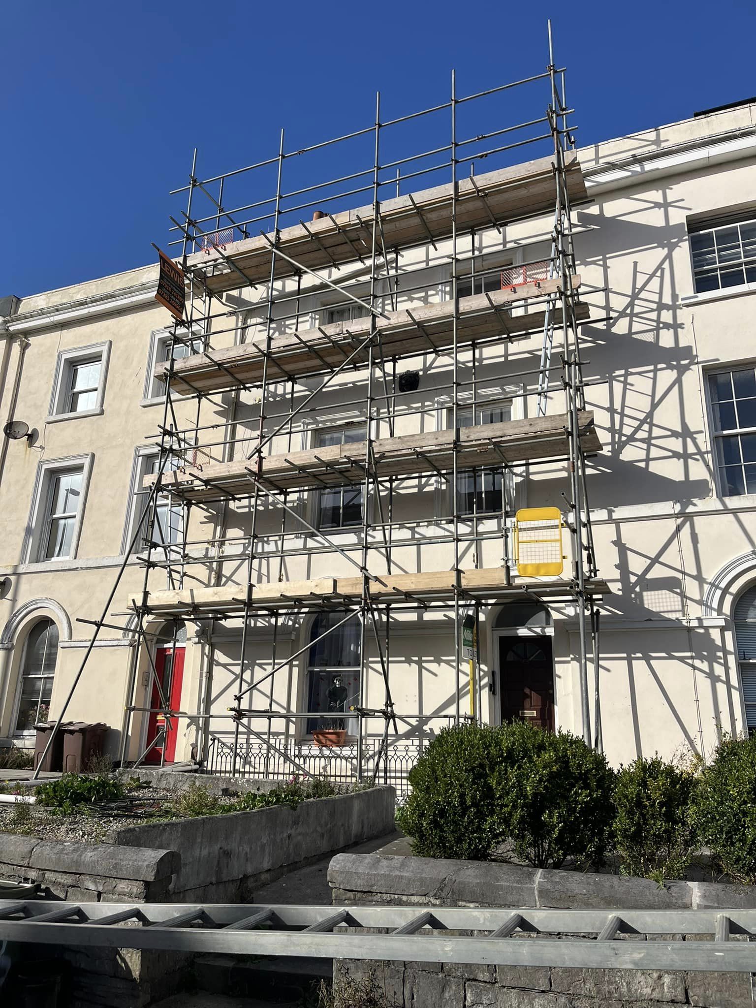 A building with scaffolding covering its facade, under a clear blue sky. The structure has several platforms, and there is a red door to the left and a black door to the right. Bushes line the front of the building.