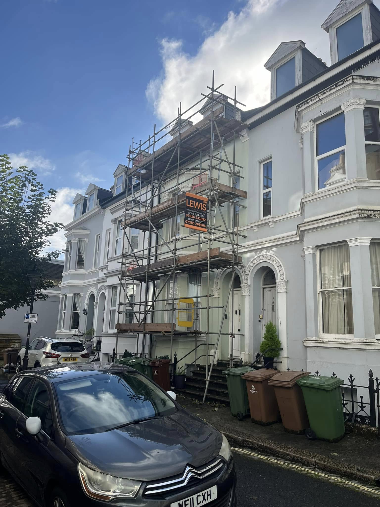 A residential street with a row of white terraced houses. One house is under renovation with scaffolding and a construction sign. Several bins are on the sidewalk, and a car is parked on the street. The sky is partly cloudy.