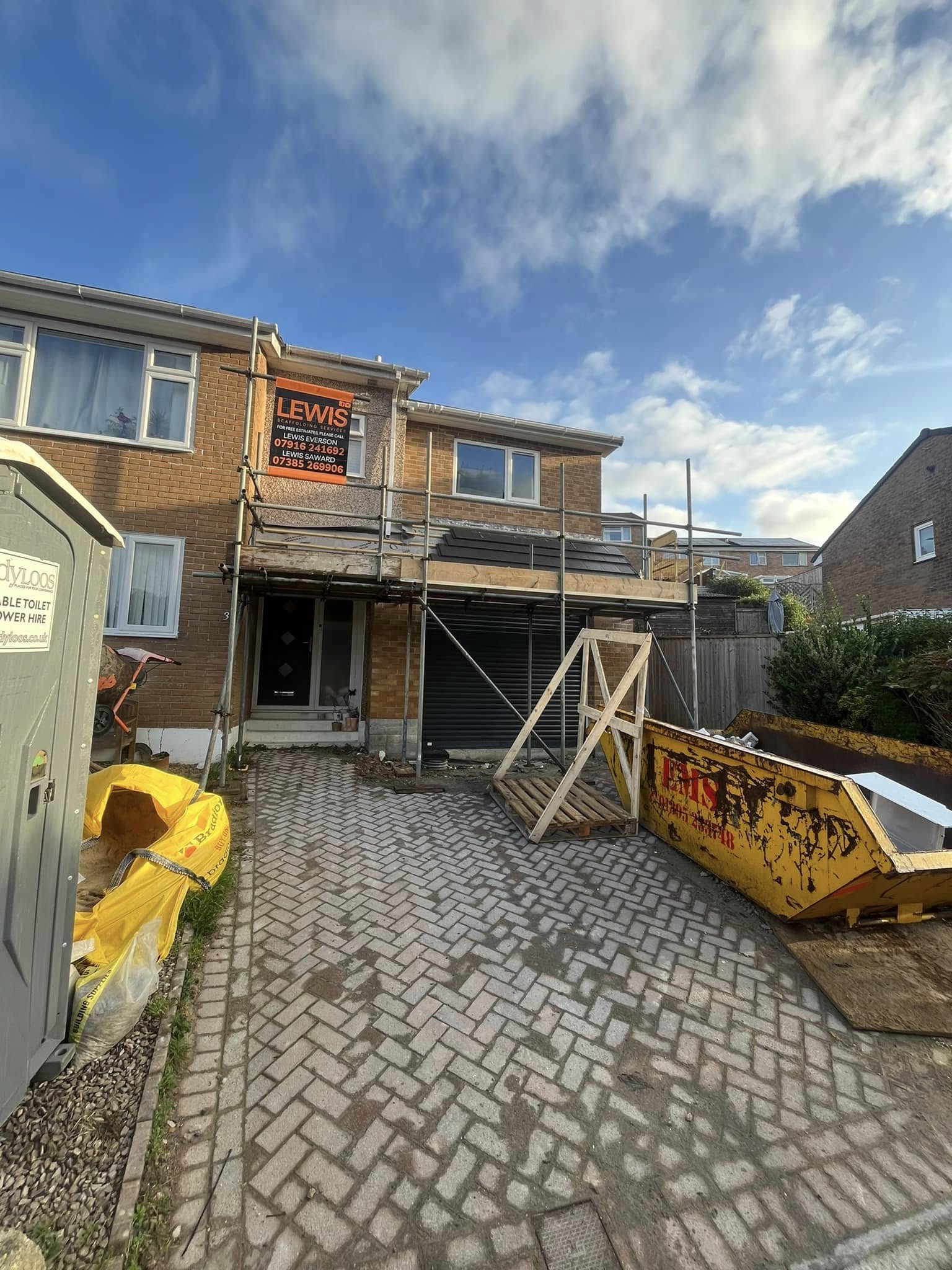 A two-story brick house under renovation with scaffolding in front. A skip filled with debris is on the driveway, which has scattered bricks. There's a portable toilet and construction materials nearby. The sky is partly cloudy.