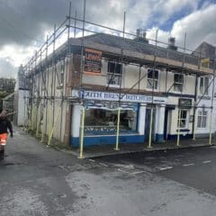 A two-story building under construction with scaffolding. The sign reads "South Brent Butchers" and features blue trim around the shopfront. A man in a safety vest walks on the left sidewalk. The sky is cloudy.