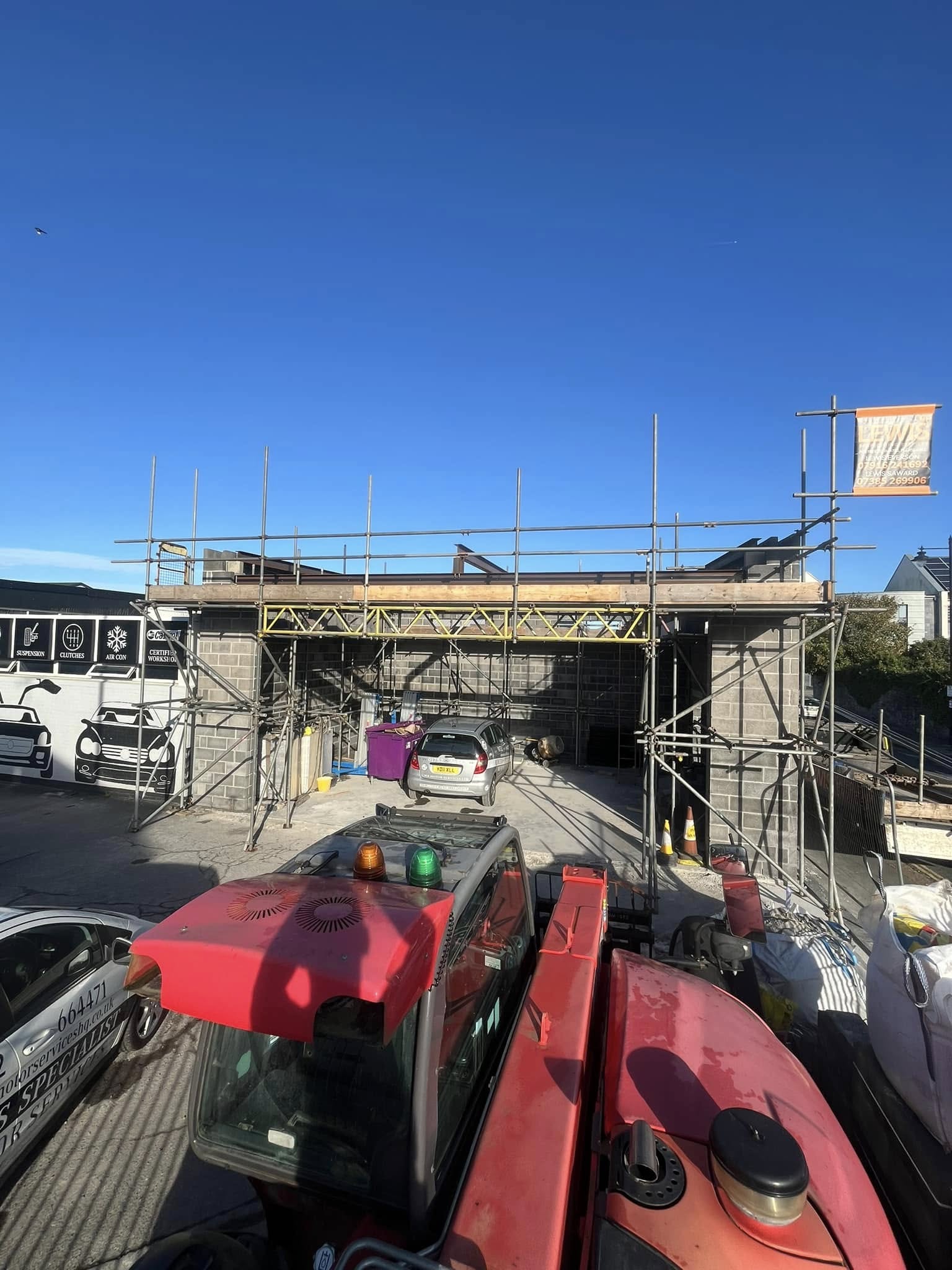 A construction site with a partially built structure and scaffolding. Several cars are parked in front, and a red vehicle, possibly construction equipment, is in the foreground. The sky is clear and blue.