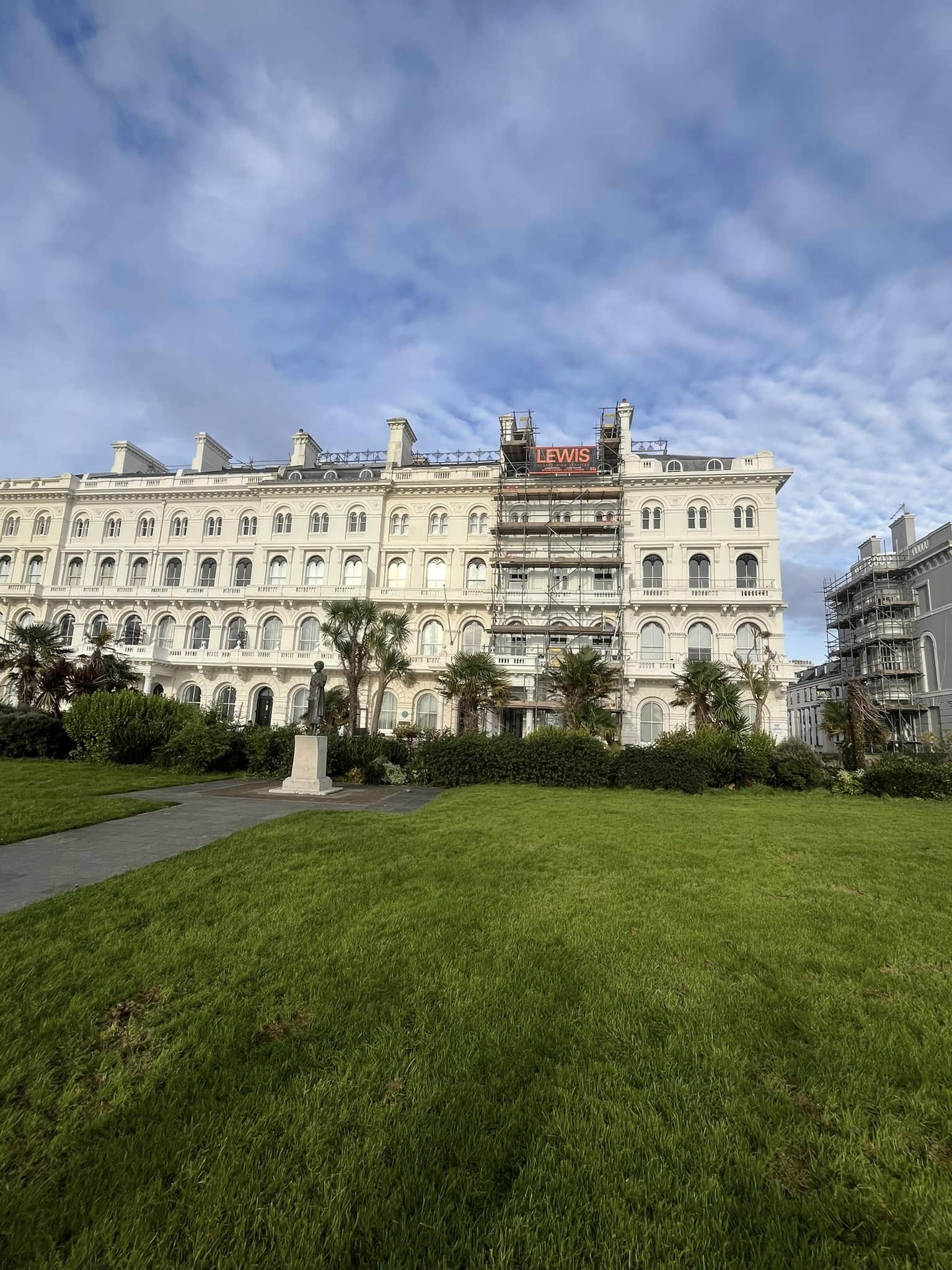 A grand white building with arched windows, partially covered by scaffolding with the name "LEWIS" visible. There is a well-maintained green lawn and trees in the foreground, set against a cloudy blue sky.