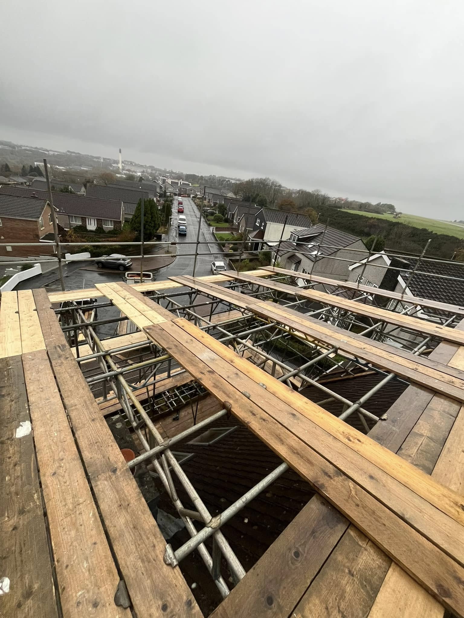 View from a rooftop under construction with wooden planks and metal scaffolding. Overcast sky with a suburban street lined with houses and leafless trees in the background.