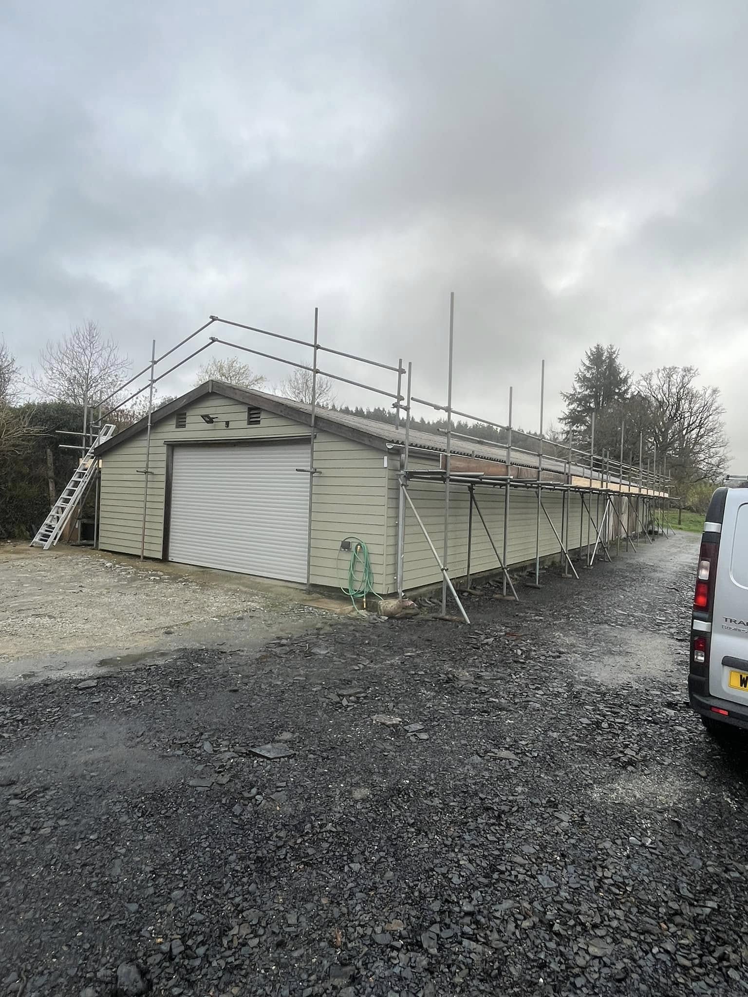 A large, light green building with a white garage door under construction, surrounded by scaffolding. A ladder leans against it, with overcast skies above. A van is parked to the right on a gravel surface. Trees are in the background.