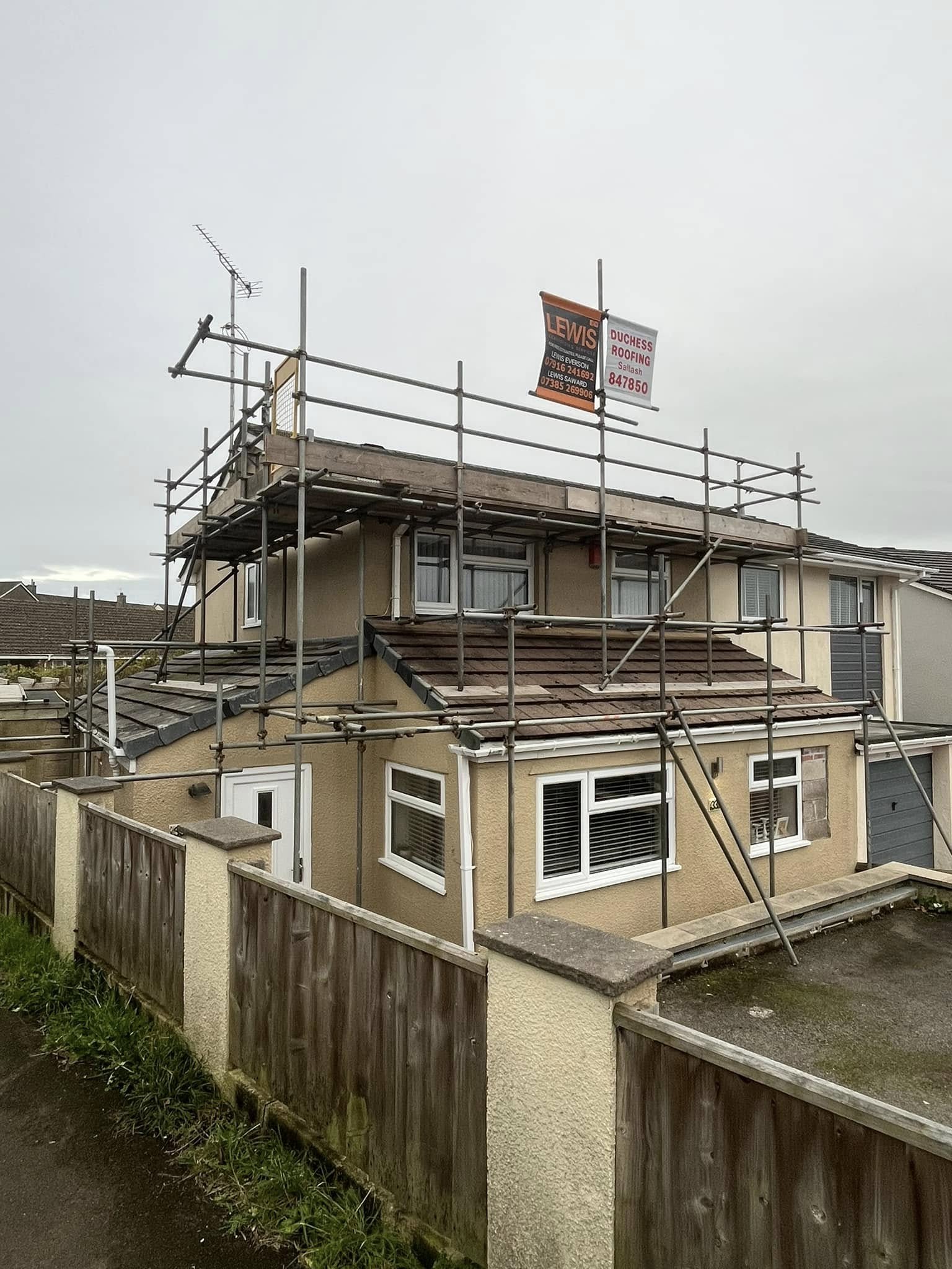 A two-story house with scaffolding around the roof, part of which is under construction. A sign for a roofing company is on the scaffolding. The house is cream-colored with brown trim, and a wooden fence surrounds the property.