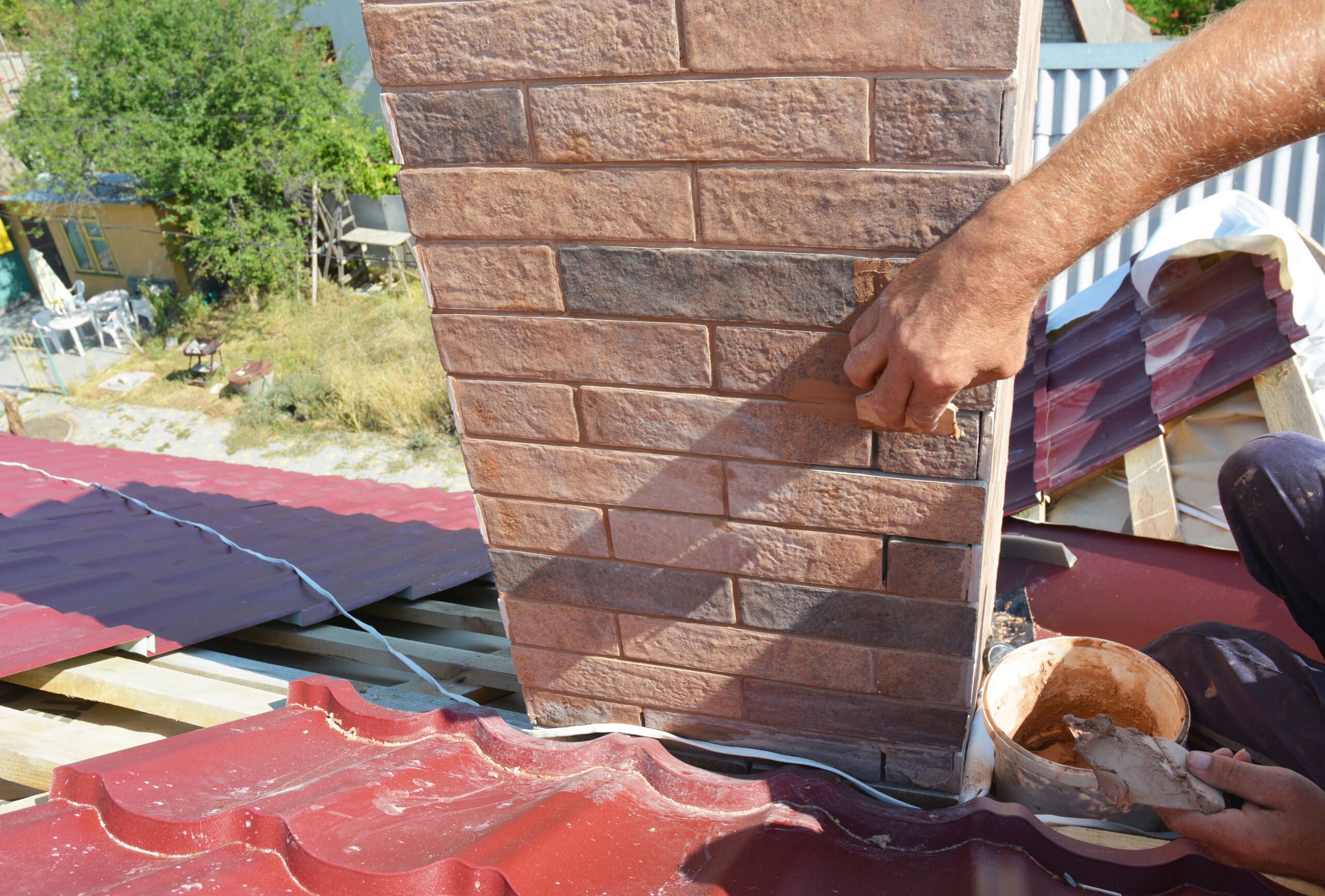 A person applies plaster to the base of a brick chimney on a red metal roof. The scene is outdoors with trees and parts of a building visible in the background.