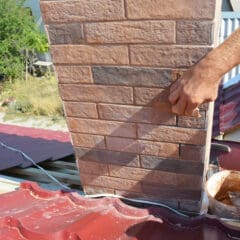 A person working on a rooftop, applying mortar to seal gaps between the chimney and red corrugated metal roofing. A bucket with mortar is on the roof beside them. The scene is set in a sunny, outdoor environment.