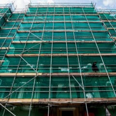 A building under construction is covered with green netting and surrounded by scaffolding. The structure reaches multiple stories high, with metal beams and wooden planks supporting the setup. The sky is partly cloudy above.