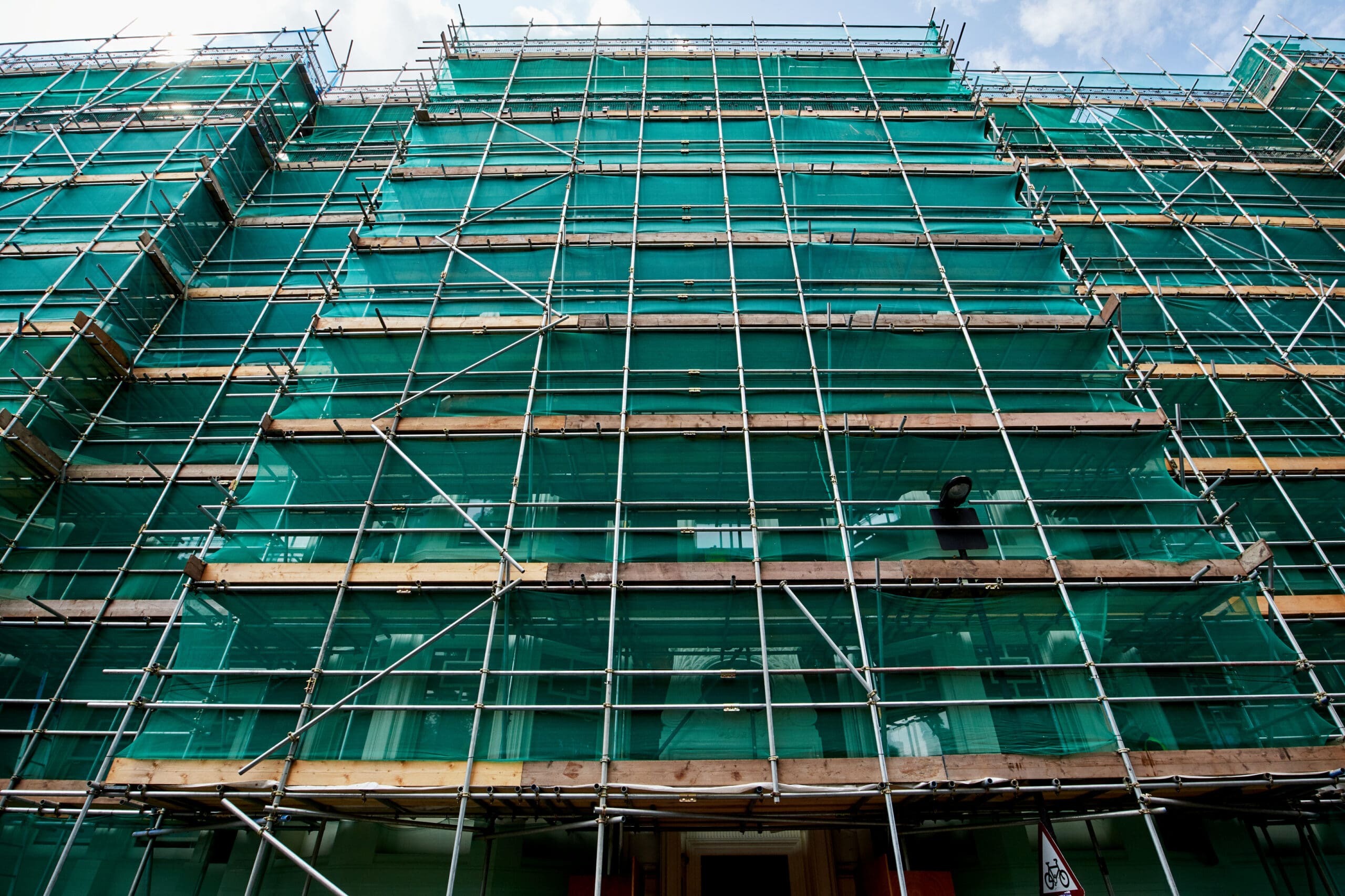 A building under construction is covered with green netting and surrounded by scaffolding. The structure reaches multiple stories high, with metal beams and wooden planks supporting the setup. The sky is partly cloudy above.