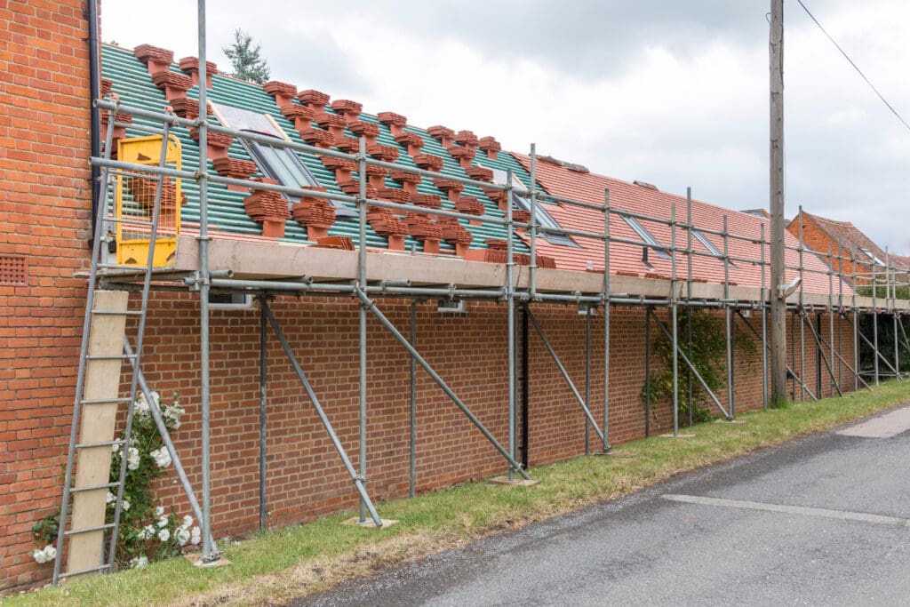 Scaffolding set up against a brick house under construction or renovation. Roof tiles are stacked in rows on the scaffold platform. A ladder leads up to the scaffold. A grass verge and road are in the foreground. Cloudy sky in the background.